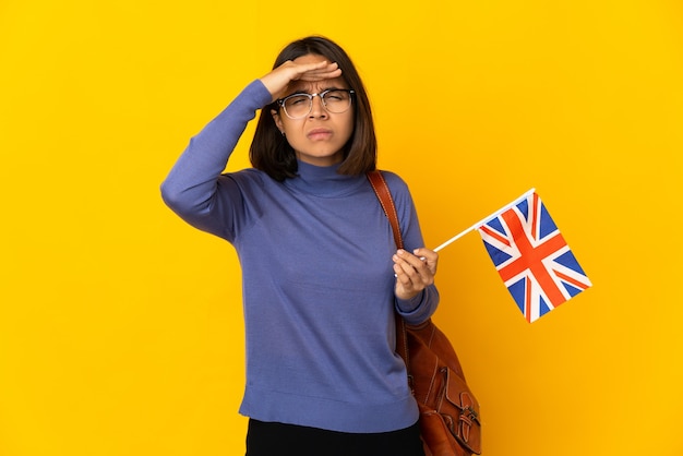 Jonge Latijnse vrouw die een vlag van het Verenigd Koninkrijk houdt die op gele achtergrond wordt geïsoleerd die ver weg met hand kijkt om iets te kijken