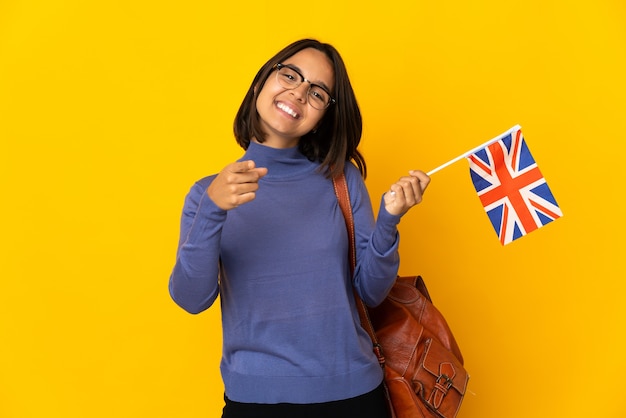 Jonge Latijnse vrouw die een vlag van het Verenigd Koninkrijk houdt die op gele achtergrond wordt geïsoleerd die voorzijde met gelukkige uitdrukking richt