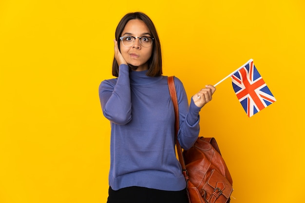 Jonge Latijnse vrouw die een vlag van het Verenigd Koninkrijk houdt die op gele gefrustreerde achtergrond wordt geïsoleerd en oren bedekt