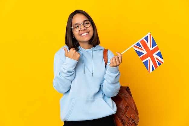 Jonge Latijnse vrouw die een vlag van het Verenigd Koninkrijk houdt die op gele muur met duim omhoog gebaar en het glimlachen wordt geïsoleerd