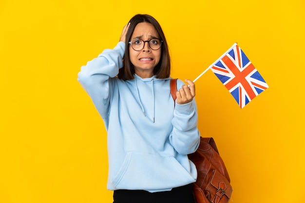 Jonge Latijnse vrouw die een vlag van het Verenigd Koninkrijk houdt die op gele muur wordt geïsoleerd die zenuwachtig gebaar doet