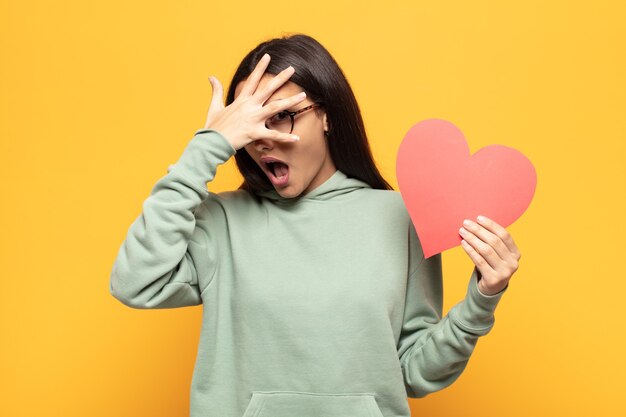 Jonge Latijnse vrouw die geschokt, bang of doodsbang kijkt, gezicht bedekt met hand en tussen vingers gluurt