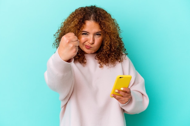 Jonge Latijnse vrouw die mobiele telefoon houdt die op blauwe achtergrond wordt geïsoleerd die vuist toont aan camera, agressieve gezichtsuitdrukking.