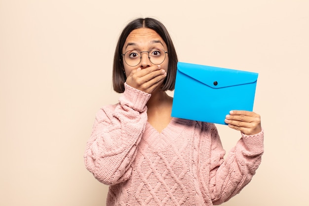 Jonge latijnse vrouw die mond bedekt met handen met een geschokte, verbaasde uitdrukking, een geheim houdt of oeps zegt saying