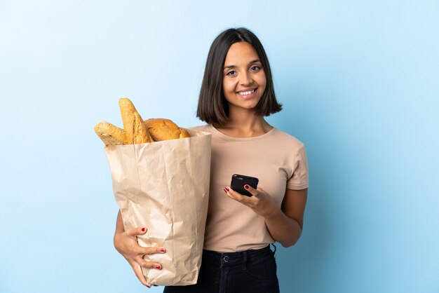 Jonge Latijnse vrouw die sommige broden kopen die op blauw worden geïsoleerd die een bericht met mobiel verzenden