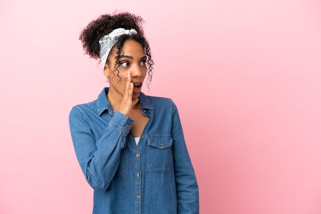 Jonge latijnse vrouw geïsoleerd op roze achtergrond die iets fluistert met een verrassingsgebaar terwijl ze naar de zijkant kijkt