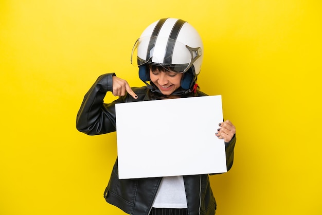 Foto jonge latijnse vrouw met een motorhelm geïsoleerd op een gele achtergrond met een leeg bordje met een gelukkige uitdrukking en erop wijzend