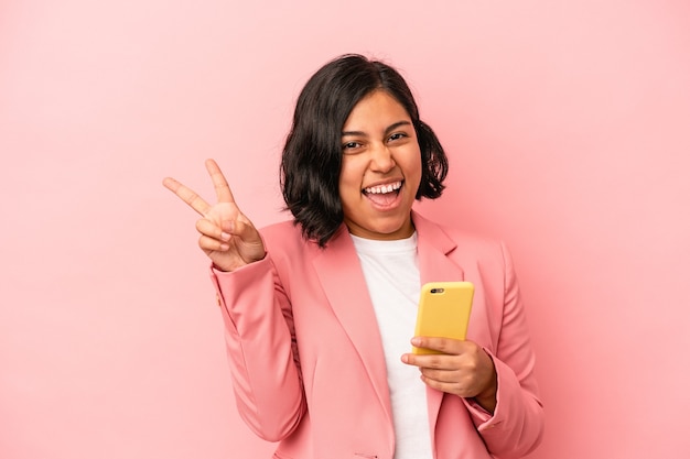 Jonge latijnse vrouw met mobiele telefoon geïsoleerd op roze achtergrond vrolijk en zorgeloos met een vredessymbool met vingers.