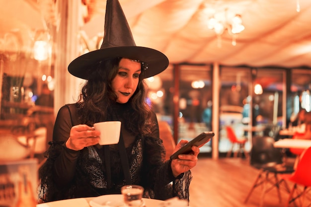 Jonge latijnse vrouw verkleed als een heks die koffie drinkt en naar de mobiele telefoon in een café kijkt