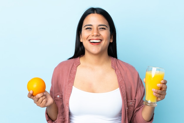 Jonge Latijnse vrouwenvrouw die op blauwe muur wordt geïsoleerd die een sinaasappel en een jus d'orange houdt