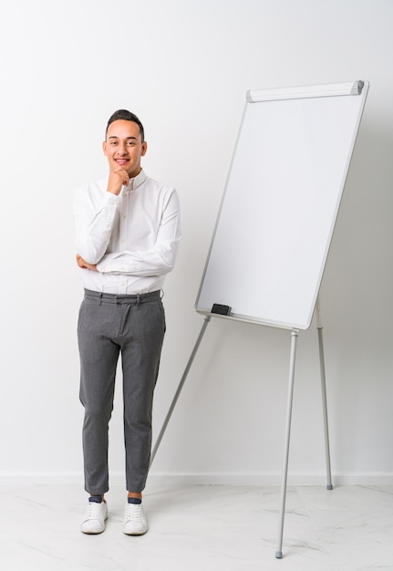 Jonge latin coaching man met een whiteboard geïsoleerd glimlachend blij en vertrouwelijk, kin met hand aanraken.