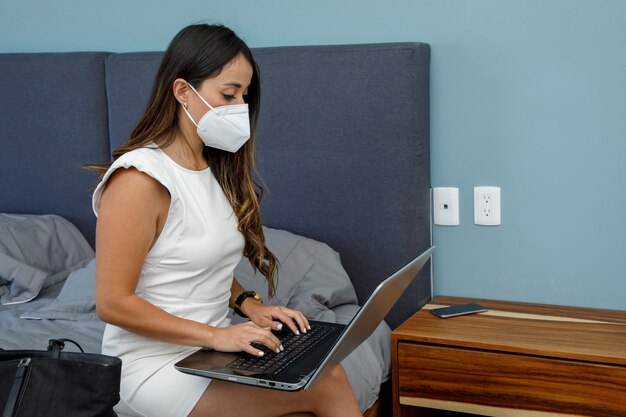 Jonge Latina zakenvrouw die een masker draagt terwijl ze op haar laptop in haar slaapkamer werkt