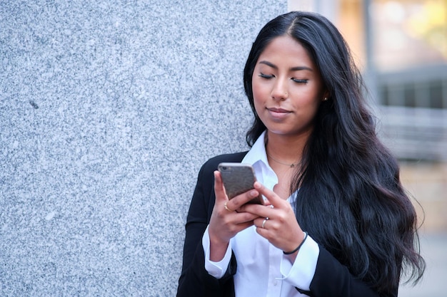 Jonge latina zakenvrouw in formele kleding die een smartphone gebruikt terwijl ze op straat staat en naar het scherm kijkt.