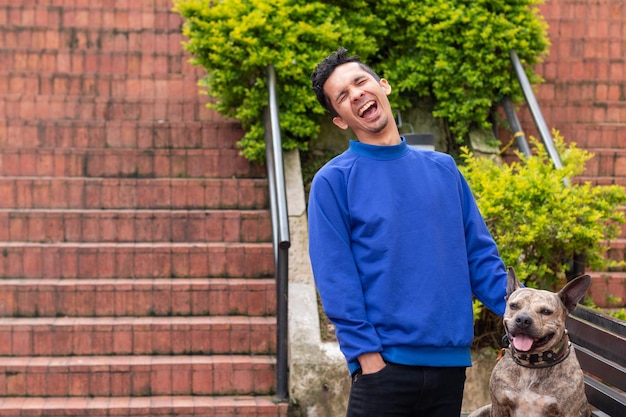 Jonge Latino man met blauwe kleren die luid lacht naast zijn huisdier pitbull