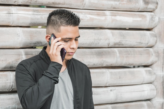 Jonge latino universiteitsstudent praten op zijn mobiele telefoon met een serieus gezicht leunend op een guadua