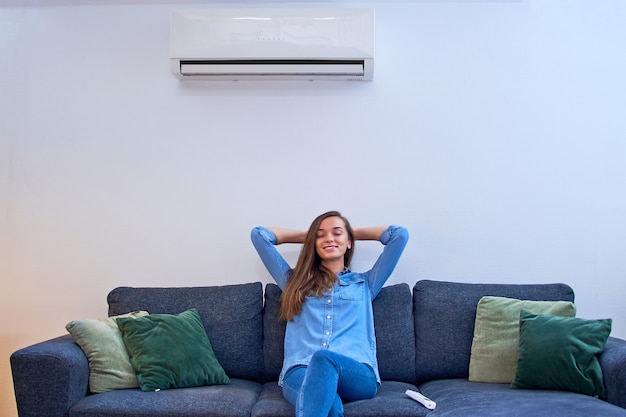 Foto jonge leuke casual rustige vrouw hand in hand achter het hoofd met gesloten ogen rustend op de comfortabele bank onder de airconditioner bij modern huis