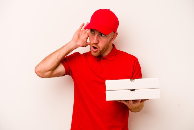 Jonge levering blanke man met pizza's geïsoleerd op een witte achtergrond probeert te luisteren naar een roddel