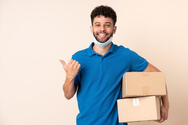 Jonge levering Marokkaanse man geïsoleerd op beige muur wijzend naar de zijkant om een product te presenteren