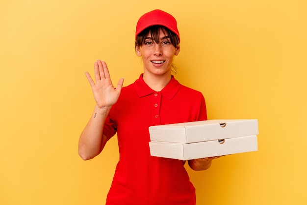 Jonge leveringsvrouw die pizza's houdt die op gele achtergrond worden geïsoleerd die vrolijk glimlachen die nummer vijf met vingers tonen.