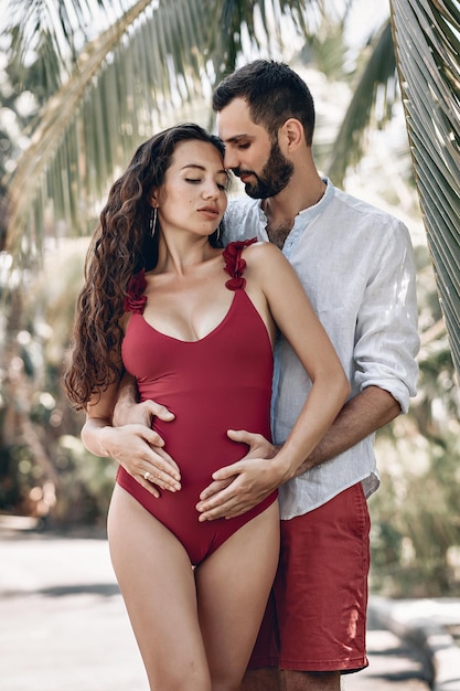 Jonge liefhebbende echtgenoot knuffelt zachtjes de buik van zijn zwangere vrouw op het tropische strand bij de palmbomen; toekomstige ouders concept.