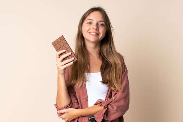 Jonge Litouwse gelukkige vrouw die op beige muur wordt geïsoleerd die een chocoladetablet neemt