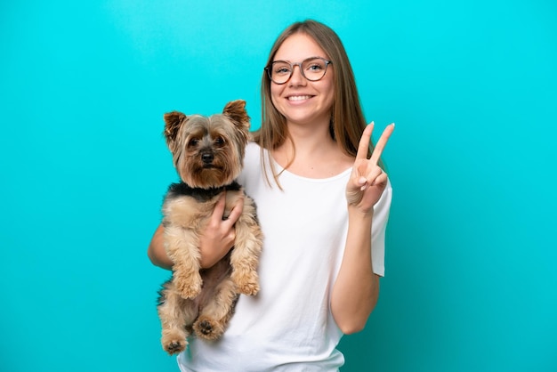 Jonge Litouwse vrouw die een hond houdt die op blauwe achtergrond wordt geïsoleerd die glimlacht en overwinningsteken toont