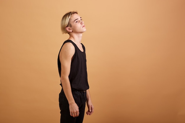 Jonge magere blonde man gekleed in een zwarte tanktop en broek poseren in de studio op de beige achtergrond.