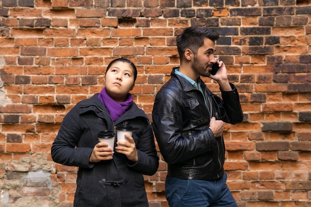 Jonge man aan de telefoon, het meisje verveelt zich en houdt twee papieren bekers met koffie op de achtergrond van een oude bakstenen muur