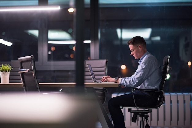 Jonge man aan het werk op laptop 's nachts in donkere kantoor. De ontwerper werkt in de latere tijd.