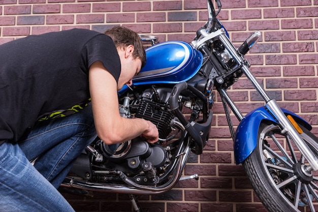 Jonge man aan het werk op zijn blauwe aangepaste motorfiets terwijl hij sommige onderdelen van dichtbij bekijkt. Gevangen voor Huis Bakstenen muur.