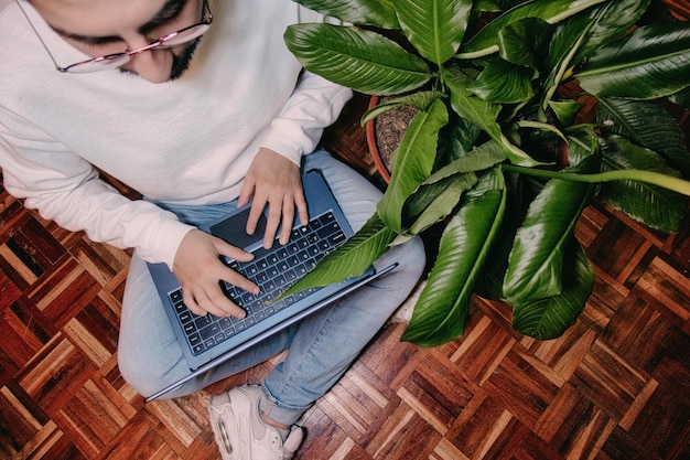 Jonge man aan het werk thuis met laptop zittend op de vloer omringd door kamerplanten