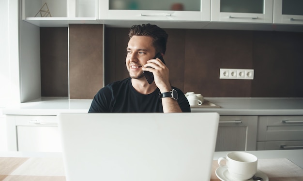 Jonge man aan het werk vanuit huis in de keuken met een laptop praten over mobiel en glimlach