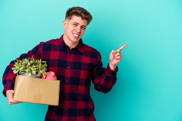 Jonge man beweegt met een doos geïsoleerd op blauwe muur glimlachend en opzij wijzend, iets laten zien op lege ruimte