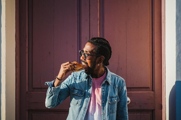 Jonge man bijt een cheeseburger op straat Het is een zonnige dag en hij heeft plezier