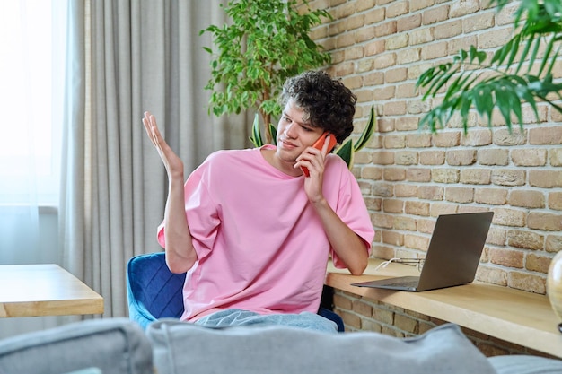 Jonge man die aan de telefoon praat en thuis aan het bureau zit met een laptop