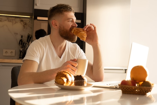 Jonge man die croissant eet en thee of koffie drinkt bij het ontbijt. doordachte europese bebaarde man zit aan tafel met eten en laptop. interieur van keuken in modern appartement. zonnige ochtendtijd