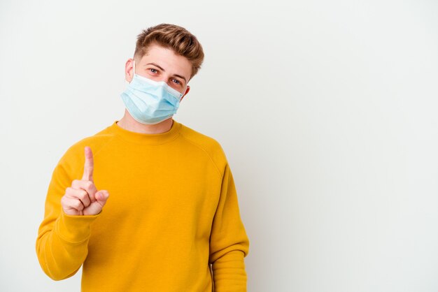 Jonge man die een masker voor coronavirus draagt dat op witte muur wordt geïsoleerd die nummer één met vinger toont.