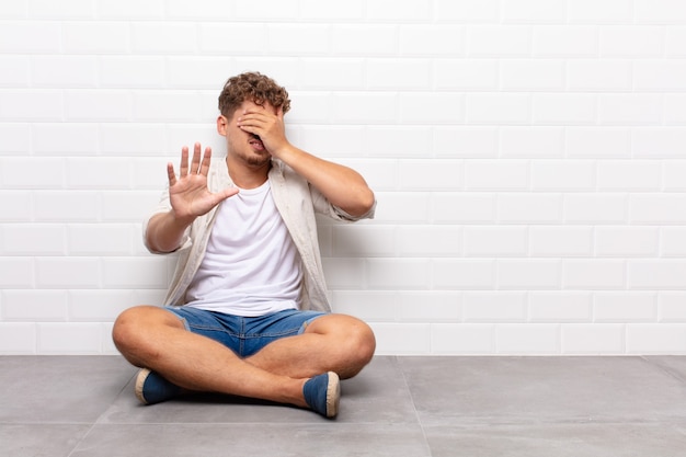 Jonge man die gezicht bedekt met hand en andere hand naar voren zet om te stoppen, foto's of afbeeldingen weigeren