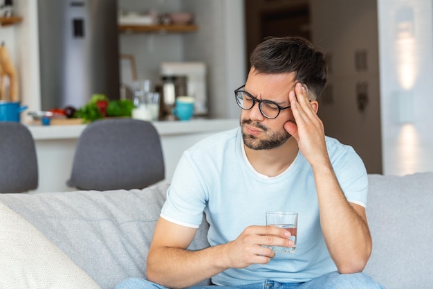 Jonge man die lijdt aan sterke hoofdpijn of migraine zit met een glas water op de bank millennial man voelt dronkenschap en pijn aanraken hoofdpijn ochtend na kater concept