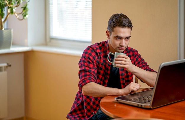 Jonge man die op een laptop werkt, zit in de buurt van een salontafel met een kop hete thee Werkplek bij het raam Werk thuis