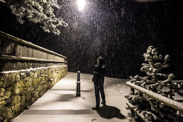 Jonge man die 's nachts naast een lantaarnpaal op een besneeuwde weg staat terwijl het sneeuwt