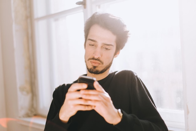 Jonge man die thuis bij het raam aan de telefoon werkt