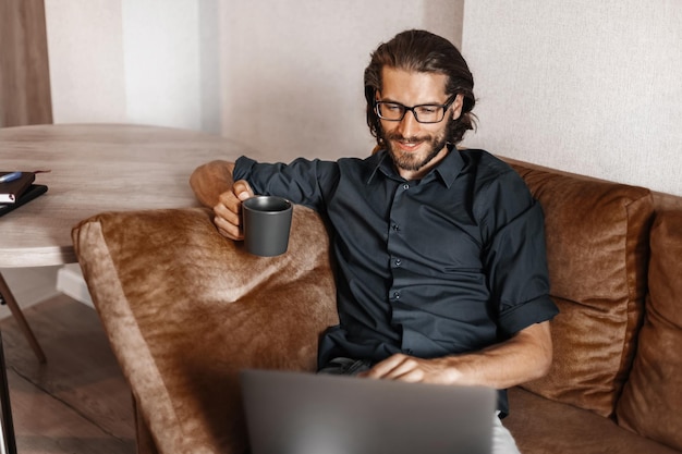 Jonge man die thuis op de bank zit, op de laptop werkt en een mok koffie vasthoudt