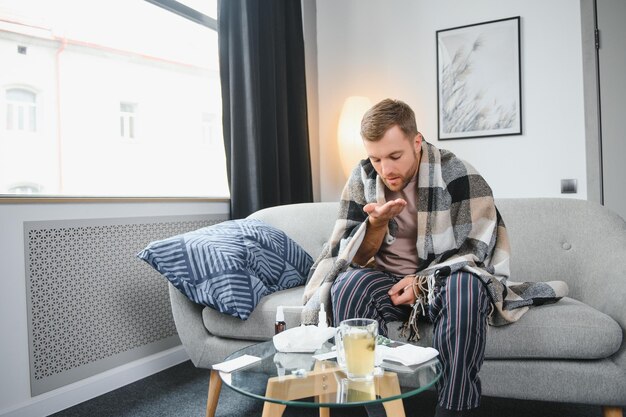 Jonge man die thuis verkouden is Schot van een jonge man die op zijn bed zit terwijl hij zich thuis onwel voelt Proberen warm te blijven Zieke man