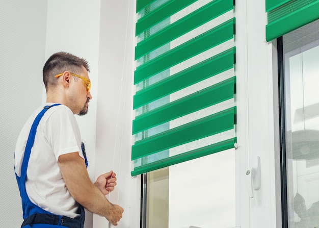 Jonge man die voor een helder raam in de kamer staat en de groene gordijnen van de kleurenblinden wit aanpast