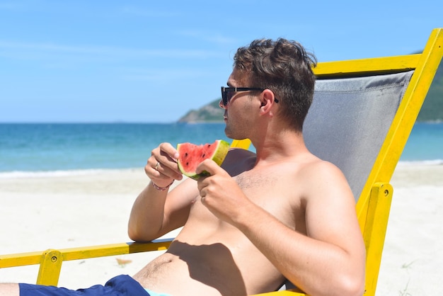 Jonge man die watermeloen eet zittend op een ligstoel op een strand in Zuid-Chinese Zee in Vietnam