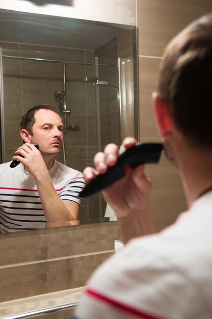 Foto jonge man die zich in de spiegel reflecteert terwijl hij zijn baard in de badkamer snijdt