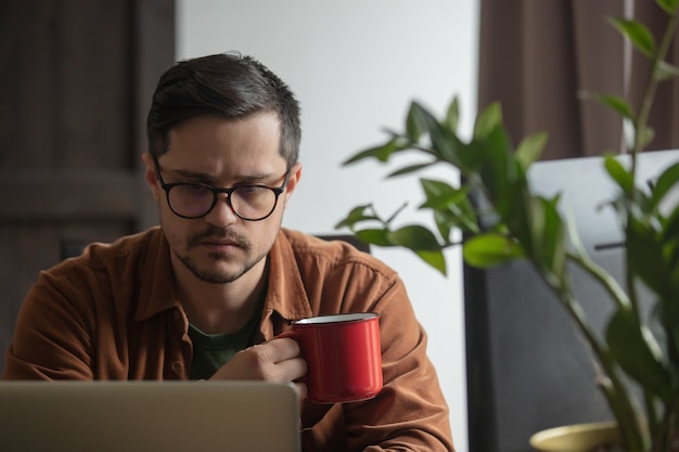 Jonge man drinkt een kopje koffie en wordt thuis wakker via laptop