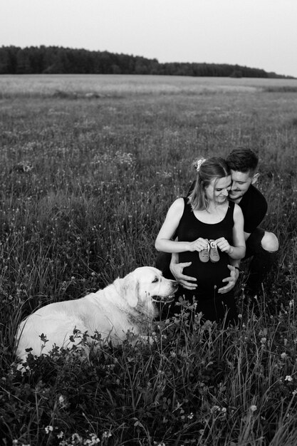 Jonge man drukt tederheid en zorg uit voor zijn zwangere vrouw, die kinderschoenen in haar handen houdt, hun slimste hond ligt naast haar. Zwart-wit foto. Grappige momenten. Nieuw leven.