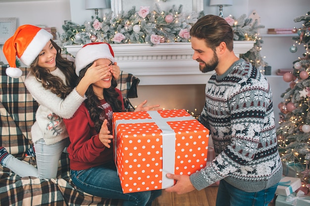 Jonge man en vrouw die grote doos heden houden. Kind houdt de ogen van haar moeder dicht. Man kijkt naar vrouw. Familie draagt feestelijke kleding.